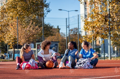 Preview: Diverse group of young woman sitting on court resting afrer playing basketball outdoors.