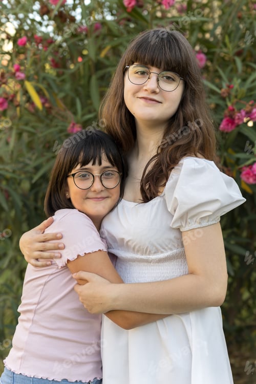Preview: Two sisters wearing glasses hug each other affectionately, smiling at the camera