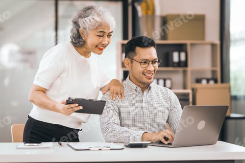 Preview: An older mature woman shows something to asian man on laptop. smile and work together in office.