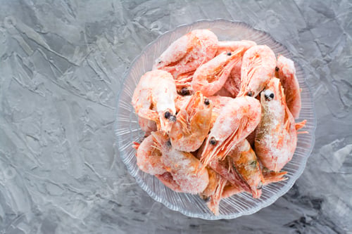 Preview: Group of frozen shrimp in a glass bowl on a gray table. Top view
