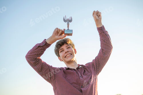 Preview: young person winning a competition, raised hands with a trophy figure against the sky with copy spac
