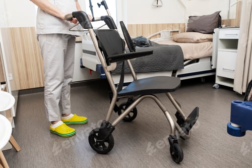 Vorschau: Einem Patienten in einer Krankenstation helfen, ins Bett zu gehen. Eine Krankenschwester hält einen Rollstuhl am Griff.