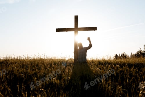 Preview: Male standing near a handmade wooden cross with his hand up towards the sky praying