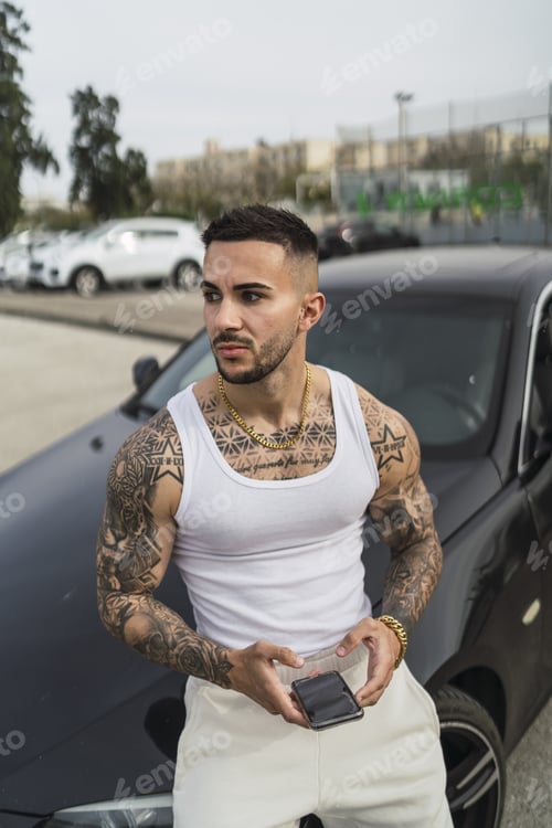 Preview: Vertical shot of a sexy tattooed male holding his phone leaning on his car