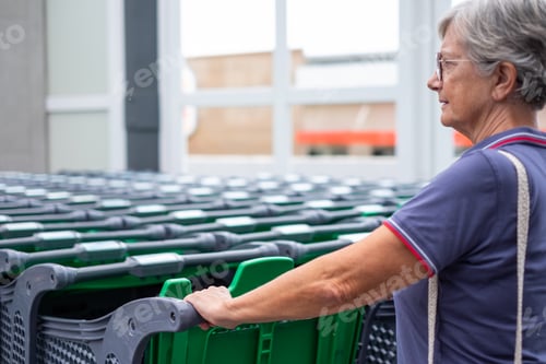 Preview: Elderly woman near a group of empty green shopping carts outside the supermarket