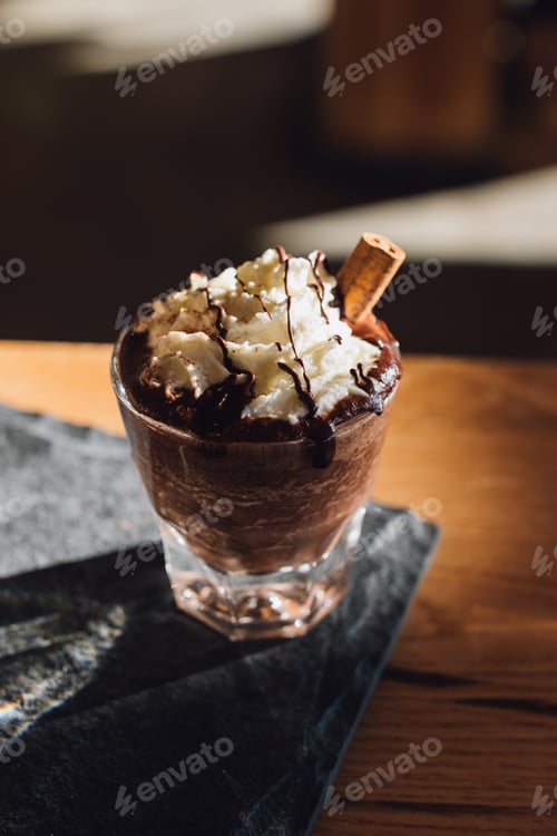 Preview: Hot chocolate topped with whipped cream, chocolate drizzle, cinnamon stick on wood table