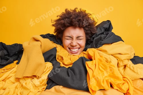 Preview: Positive woman with curly hair covered by stack of clothing does laundry at home smiles broadly keep