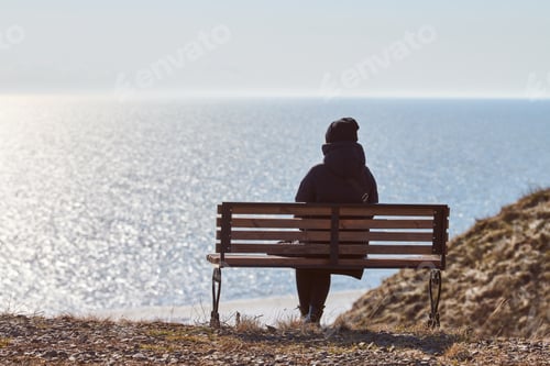 Preview: Single girl in a black jacket and hat sitting on bench at cliff at front of sea peaceful quiet place