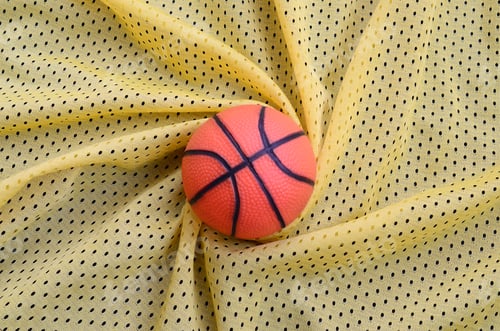Preview: Small orange rubber basketball lies on a yellow sport jersey clothing fabric texture