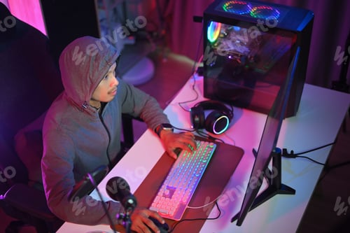 Preview: Professional gamer in hoodie playing online video game at home with neon decorated light