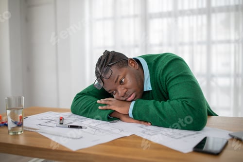 Preview: Frustrated young man with glasses rests his head on work desk in bright room