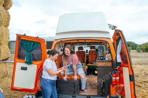 Preview: Two women looking at phone beside camper van and hay bales.