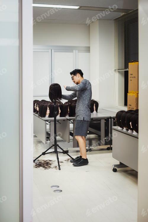 Preview: Bearded man wearing glasses standing indoors, cutting hair of brown wig on mannequin head.