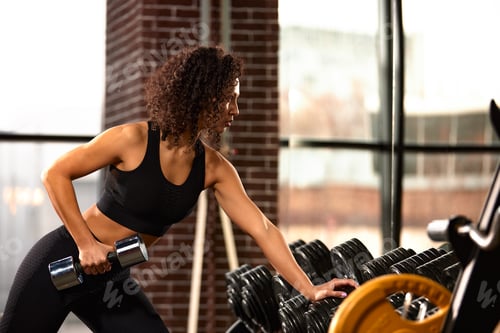 Preview: An African American woman trains with dumbbells in the gym in front of a mirror, doing a workout on