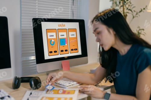 Preview: Asian woman designs a mobile app, focusing on details, in a cluttered office. Screens display code a