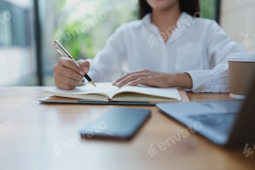 Preview: Female professional writing notes in a notebook with a pen, surrounded by a laptop and coffee, creat