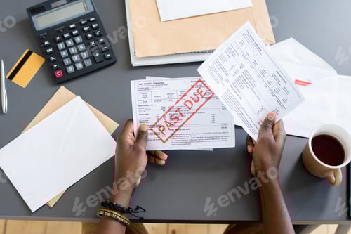 Preview: Hands of young African male holding unpaid bill over other papers and invoices