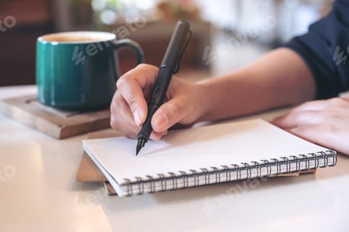 Preview: Closeup image of a woman's hand writing on blank notebook with coffee cup on table in cafe