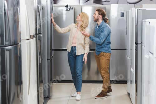 Preview: smiling boyfriend pointing with hand and girlfriend touching fridge in home appliance store