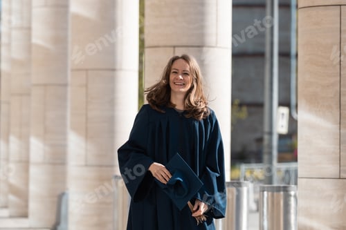 Preview: White middle aged woman in blue gown smiling and holding her cap in hands. Graduation. Diversity.