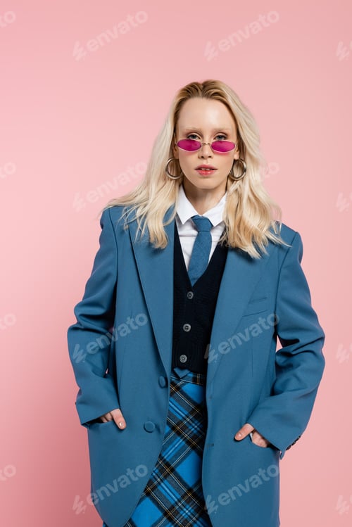Preview: blonde woman in blue blazer with tie and sunglasses posing with hands in pockets isolated on pink