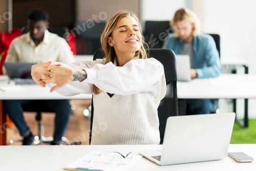 Preview: Young, happy businesswoman stretching at her desk in busy office
