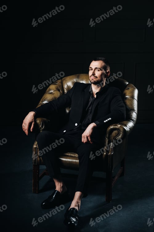 Preview: portrait of an elegant young European man in a suit sitting on a chair in a dark interior