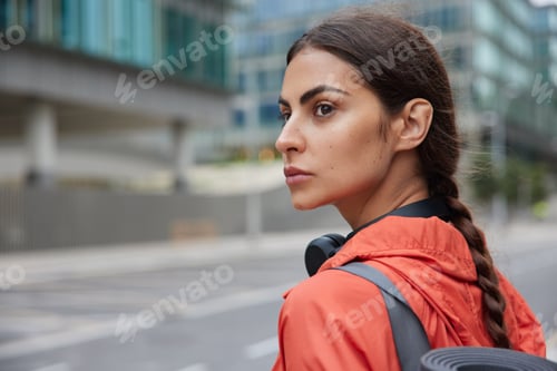 Preview: Serious brunette woman with pigtail wears jacket headphones around neck looks away strolls in big ci