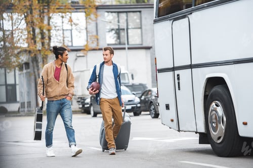 Preview: adult man with rugby ball carrying travel bag while his mixed race male friend walking near bus at