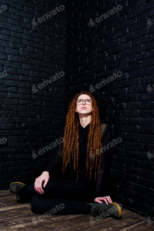 Preview: Studio shoot of girl in black with dreads, at glasses and hat on brick background.