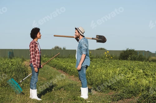 Preview: side view of happy interracial farmers with shovel and rakes looking at each other on farmland