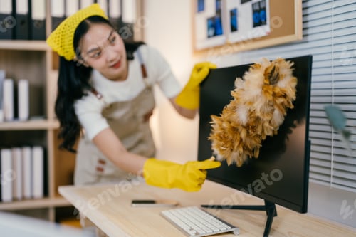 Preview: Young smiling maid wearing yellow gloves is carefully dusting a computer screen with a feather duste