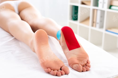 Preview: Cropped view of young woman with kinesiology tapes on foot lying on massage couch in clinic