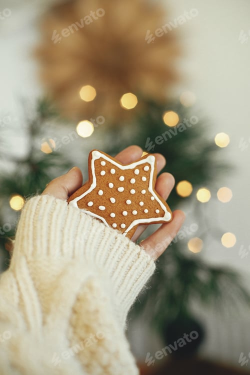 Preview: Hand in cozy sweater holding gingerbread cookie star on moody background with lights. Happy Holidays
