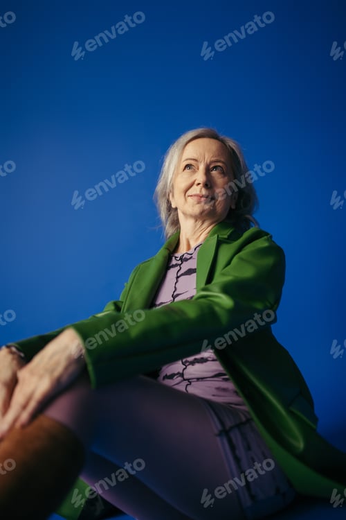 Preview: grey haired woman in green leather jacket and purple dress smiling and looking away on blue