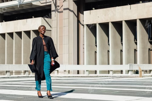 Preview: African American woman walking on zebra crosswalk at campus, wearing gown, holding cap, copy space