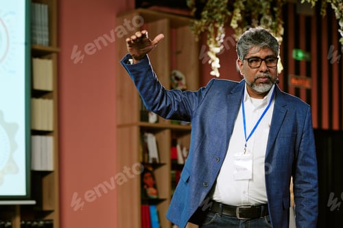 Preview: Middle Aged South Asian Man Giving Presentation in Front of Bookshelf