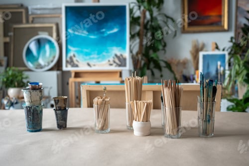 Preview: Pile of wooden sticks, brushes distributed in cups standing on table against of art studio workshop