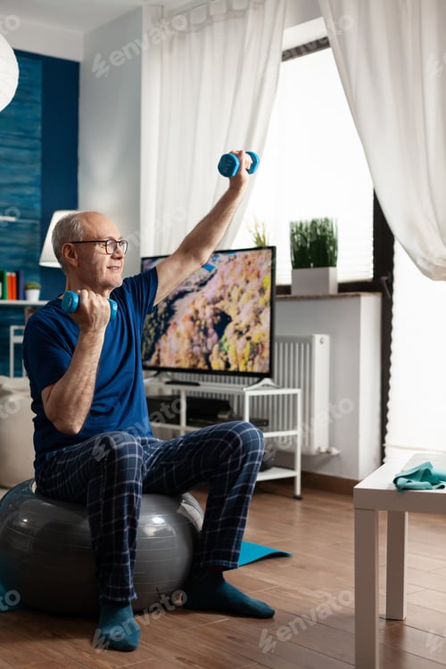 Preview: Retirement senior man sitting on aerobics swiss ball raising hand streching arm muscle