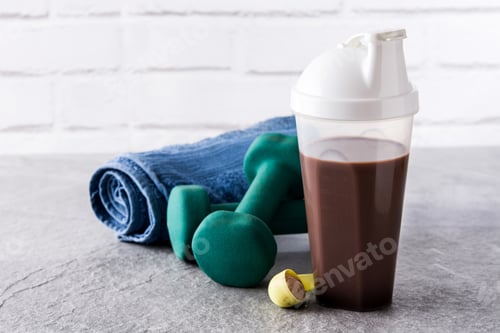 Preview: Chocolate protein shake, dumbbells and towel resting on gray surface