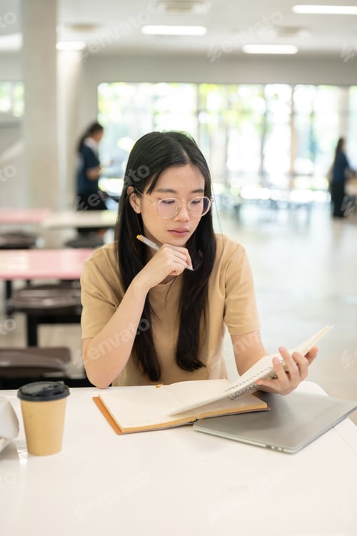 Preview: Glasses woman college student holding pen reading book over laptop sitting at table in the building.