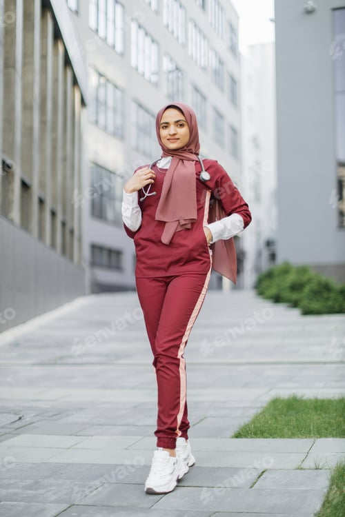 Vista previa: Young Muslim lady doctor, wearing red medical scrubs and treaditional hijab, posing outdoors