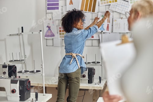 Preview: African-American fashion designer puts dresses models on wall looking at mature colleague