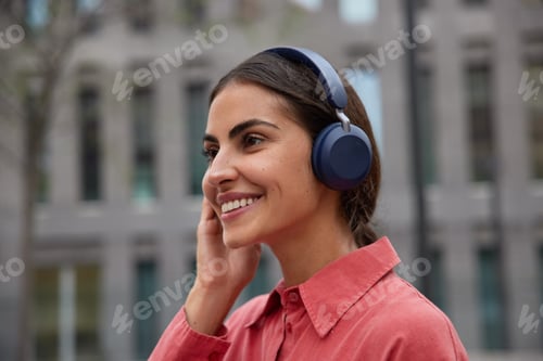Preview: Sideways shot of happy dark haired woman listens music in headphones outdoors smiles gently focused