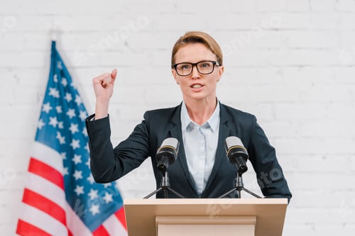 Preview: emotional speaker in glasses gesturing while talking near microphones