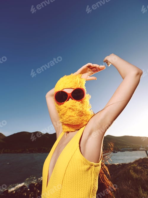 Preview: Woman in yellow dress and sunglasses with arms raised standing by the ocean, mountain range in