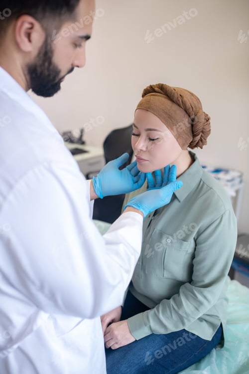 Preview: Tall doctor in blue gloves looking concentrated while palping lymph nodes of his patient