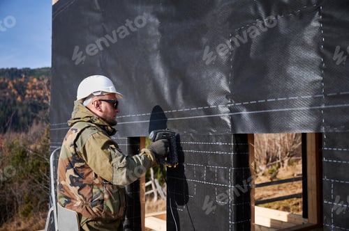 Preview: Male builder installing waterproof membrane on wall of future wooden frame house.