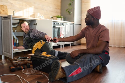 Preview: Young black man in workwear passing handtool to mature colleague