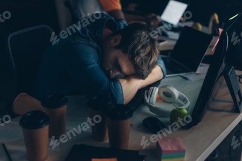Preview: young, exhausted programmer sleeping at workplace at night in office